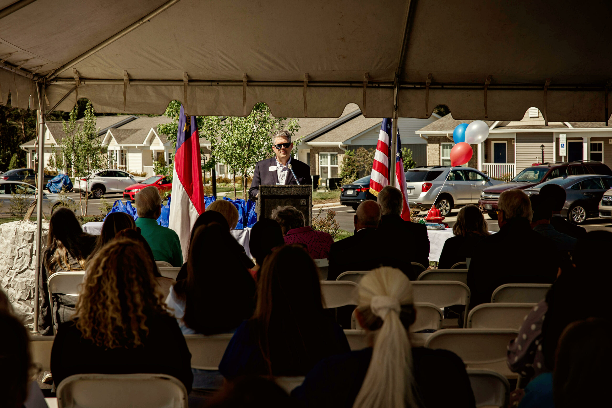 a man is at a podium in front of a crowd. They are outdoors under an event tent, and flags are on either side of the podium.