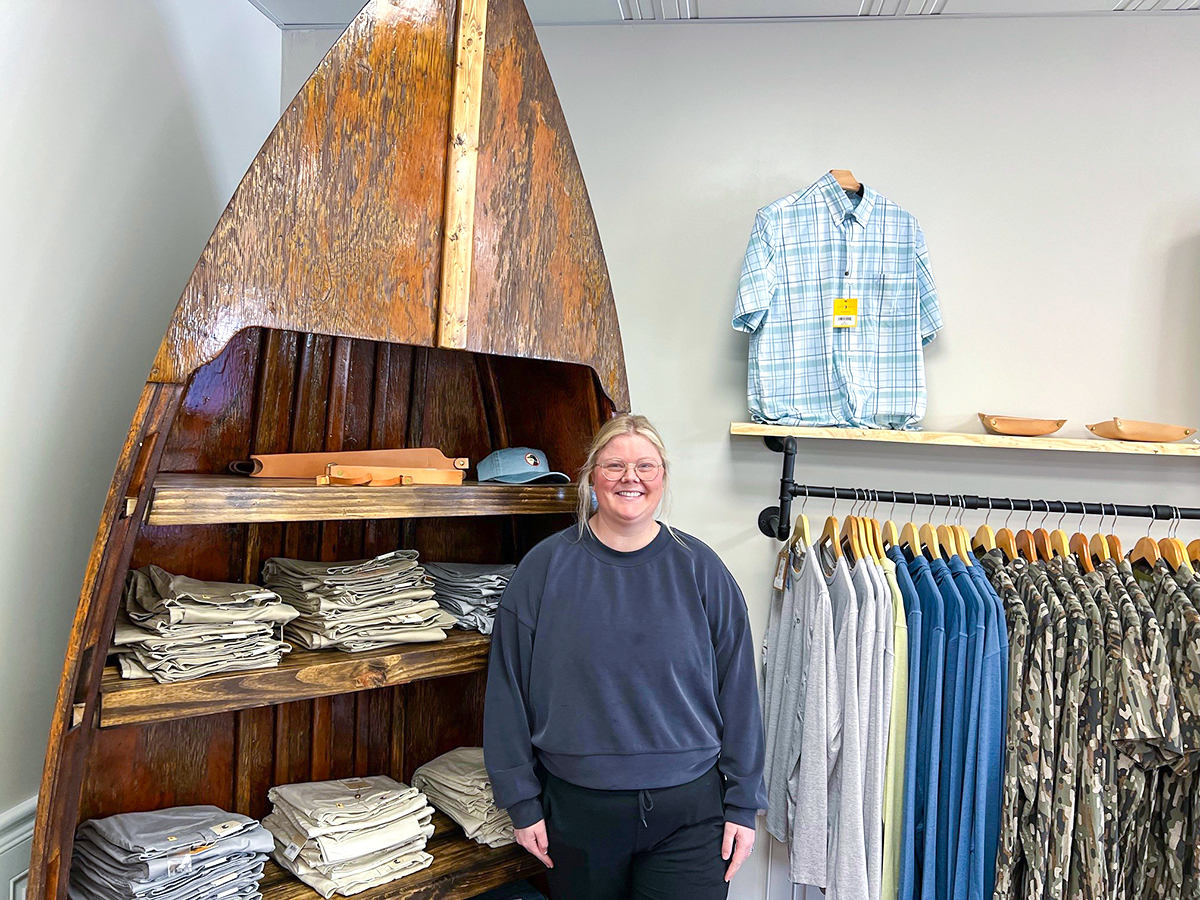 Owner of store, Barfield’s Mercantile standing in front of restored wooden Jon Board holding clothes on display and a rack of shirts hanging up next to her.