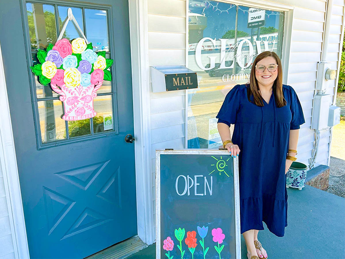 Owner, Amanda Winters standing in front of an open sign to her shop, Glow Therapy Clothing.
