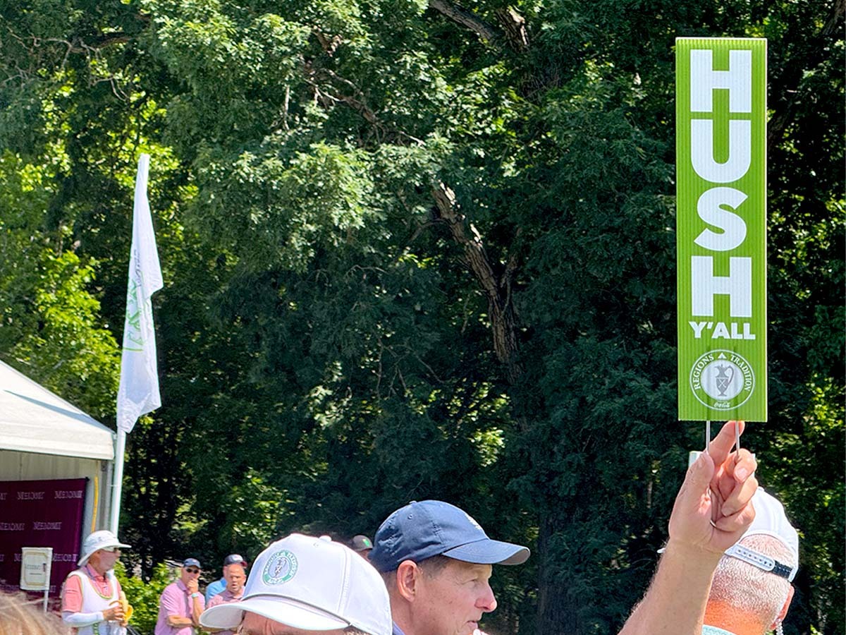 A view of the tops of peoples heads in a crowd as a man holds up a sign that says, "Hush Y'all."