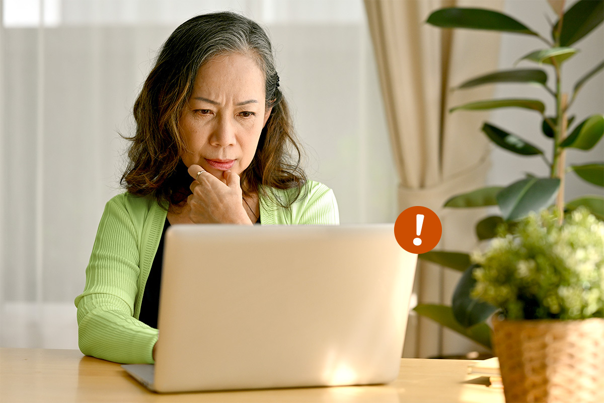 Asian American senior woman sitting at a computer with a concerned look on her face. An illustrated red exclamation mark if coming off the top right of the computer.