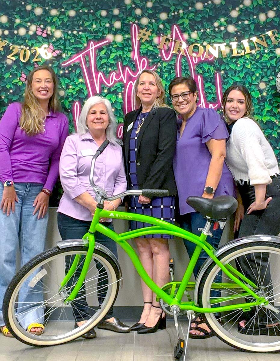 Group photo of Regions assiciates posing with a green bike in front of a floral, "Thank you" backdrop. 